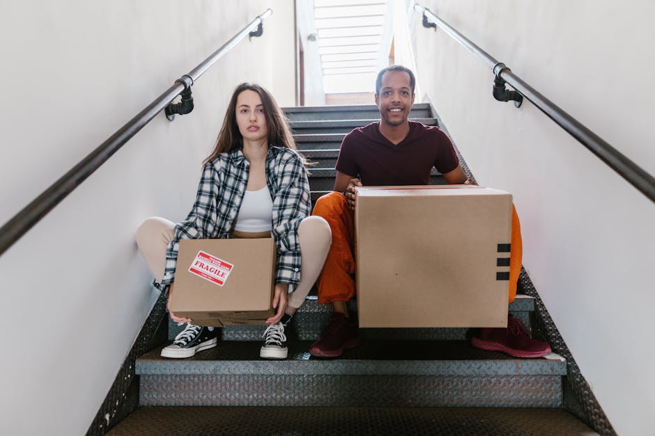 A young woman with long brown hair, wearing a black and white checkered shirt and casual sneakers, sits on a staircase holding a small cardboard box labeled 'Fragile' in red and white. Beside her, a young man with short hair, wearing a maroon T-shirt and orange pants, smiles while carrying a larger cardboard box. Both individuals are positioned on a staircase inside a building, with metal handrails on either side, leading up to a window with horizontal blinds, allowing natural light to illuminate the scene. The stairs are made of dark material with a textured surface, and the walls of the interior are light-colored. The loading process appears to be part of a home relocation or furniture transport operation, with the individuals preparing to move boxes as part of the packing and moving stage. This image exemplifies a typical step in house removals involving loading boxes on stairs before transferring them to a vehicle, which Man and Van Brentford offers as part of their relocation services.