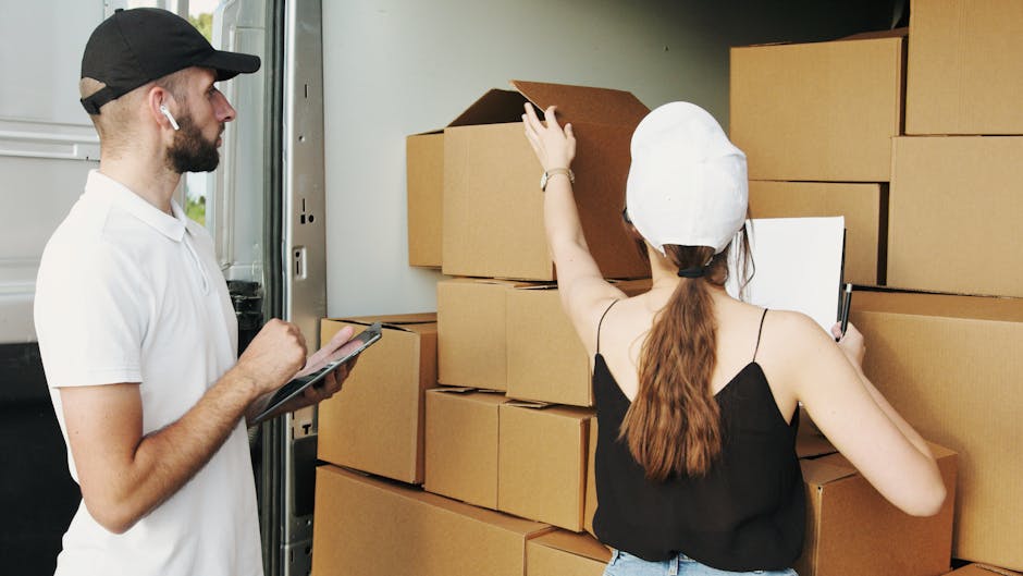 A young woman with long brown hair, wearing a black and white checkered shirt and casual sneakers, sits on a staircase holding a small cardboard box labeled 'Fragile' in red and white. Beside her, a young man with short hair, wearing a maroon T-shirt and orange pants, smiles while carrying a larger cardboard box. Both individuals are positioned on a staircase inside a building, with metal handrails on either side, leading up to a window with horizontal blinds, allowing natural light to illuminate the scene. The stairs are made of dark material with a textured surface, and the walls of the interior are light-colored. The loading process appears to be part of a home relocation or furniture transport operation, with the individuals preparing to move boxes as part of the packing and moving stage. This image exemplifies a typical step in house removals involving loading boxes on stairs before transferring them to a vehicle, which Man and Van Brentford offers as part of their relocation services.