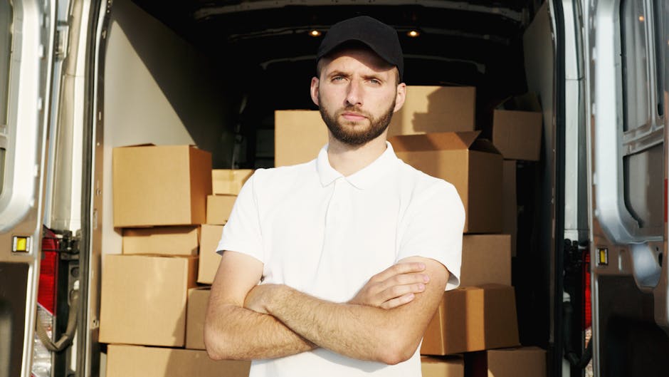 A man with a serious expression, wearing a black cap and a white polo shirt, stands with arms crossed inside the back of a moving van filled with tightly stacked cardboard boxes of various sizes. The boxes are securely packed and wrapped with plastic for protection during a house removal. The van's interior is well-lit, and the open rear doors reveal a clear pathway for loading and unloading furniture and packaging materials. The scene illustrates a professional furniture transport process as part of home relocation, with the man overseeing the packing and loading process. Man and Van Brentford’s services involve careful packing, transport, and handling of household items, ensuring efficient and safe house removals.