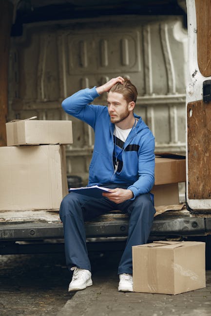 A young man with light brown hair and a beard, dressed in a blue tracksuit and white sneakers, is sitting on the edge of the open vehicle van used for home relocation. He is inside the vehicle, which has a metal interior with visible ribs and a wooden panel on the right side. Around him, there are several cardboard boxes of varying sizes, some stacked and others placed on the ground, likely filled with household items or personal belongings. The man is holding a clipboard and pen, with his left hand resting on his head as he appears to be in thought or reviewing a moving checklist. The scene is set outdoors during daylight, with natural light illuminating the area, and the van's rear door is open, providing access for loading and unloading furniture and boxes. This image effectively illustrates the packing and loading process involved in house removals, as performed by [COMPANY_NAME], highlighting the careful handling and planning necessary for successful furniture transport and moving services.