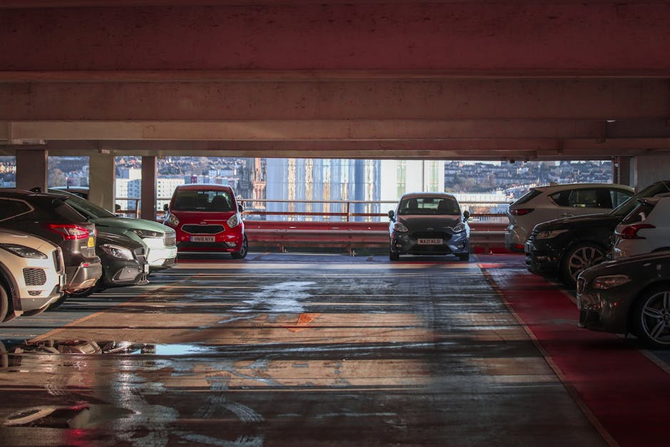 In a multi-storey parking garage, several vehicles including a red van and various cars are parked on the concrete floor, which is wet and has visible tire marks. The garage features support columns and a low ceiling painted in a reddish hue. Natural light and cityscape views are visible through the open side of the structure, showing tall buildings in the distance. The scene depicts a typical arrangement of parked vehicles, with some positioned near a designated loading area for house removals. In the context of home relocation, the image illustrates a loading process where furniture and packing materials could be transferred from the property into the vehicles. The presence of [COMPANY_NAME], such as Man and Van Brentford, would manage the logistics of furniture transport, including packing and loading inside the garage space. The parking area appears prepared for removal vehicles to load and unload household belongings efficiently, with ample space for manoeuvring large items and equipment like trolleys or straps, critical for safe furniture transport during a house move.