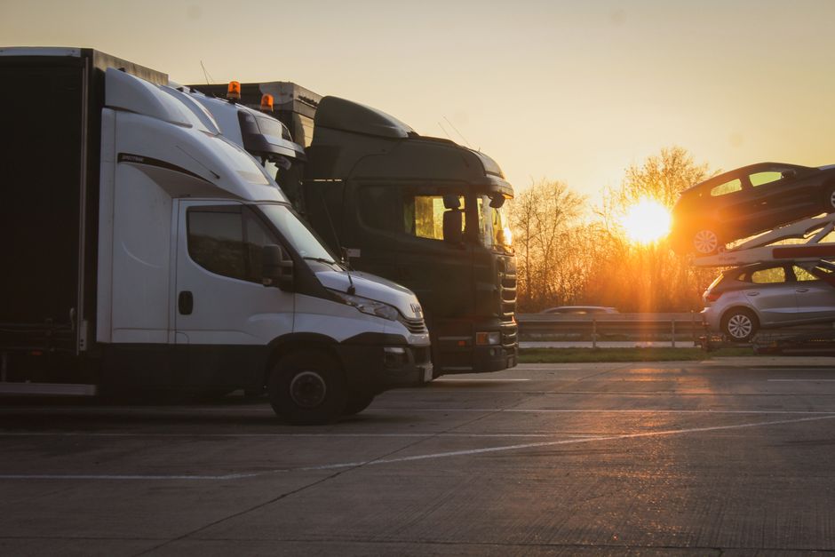 A scene at a parking lot during sunset shows two large removal trucks parked side by side, one white and one black, with an interior view of a home relocation process. The white truck is on the left, partly visible, with its side door and a portion of the cargo area showing. The black truck is positioned directly behind it. To the right, a car carrier trailer is transporting two cars stacked vertically—one dark-colored vehicle on top and a lighter one below—secured during transport. The parking area has marked spaces and a smooth concrete surface, with a few other cars visible in the background on a busy road. Leafless trees and a bright, setting sun create warm lighting, casting long shadows. Occasionally, furniture and packing materials like cardboard boxes and blankets may be seen being loaded or unloaded, supported by the visible lifting and loading actions. This scene illustrates the logistics involved in house removals, with vehicles equipped for furniture transport and packing for a home relocation, as managed by [COMPANY_NAME], in compliance with Brentford parking suspensions for removals vehicles.