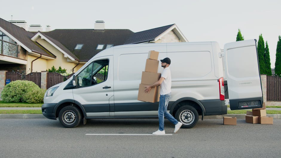 Inside the open rear of a white moving van positioned on a paved driveway, two young women with casual clothing and long hair are seated on the edge of the vehicle's floor. One woman has curly black h