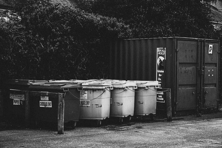 A man wearing a dark jacket and cap standing on a dirt ground near heaps of assorted household waste and recyclables, including plastic pipes, metal frames, electronic components, cardboard boxes, and
