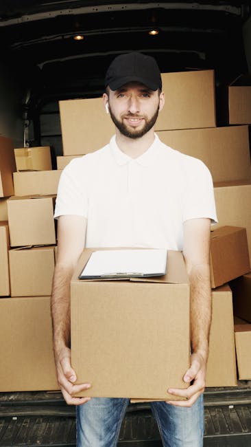 A young man with a beard, wearing a black cap, white t-shirt, and jeans, stands inside the open back of a black van used for home relocation. He is holding a medium-sized cardboard box in front of him