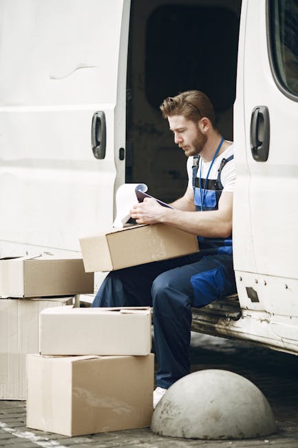 A young man with a beard, wearing a black cap, white t-shirt, and jeans, stands inside the open back of a black van used for home relocation. He is holding a medium-sized cardboard box in front of him