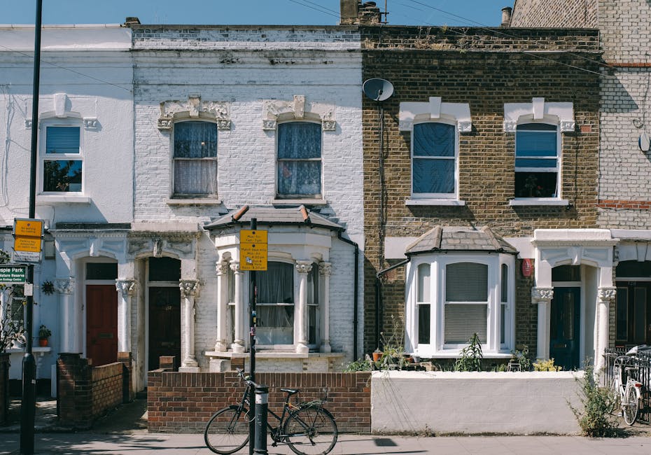 The image depicts the front façade of two adjoining terraced houses with brick and painted exteriors, featuring bay windows on the ground floor and standard windows above. The house on the left has a white painted façade with decorative stonework around the bay window and a brown front door, while the house on the right has a darker brick façade with white trim around its bay window and a black door. In front of the houses, a low brick wall encloses small front gardens, with some potted plants visible near the entrances. A black bicycle is parked against a lamppost situated on the pavement in front of the houses, which are positioned on a residential street. Two yellow parking restriction signs are mounted on the lamppost. The setting appears well-lit with natural daylight, and the overall scene is indicative of an urban neighborhood typical for home relocation or furniture transport activities, with no objects or people actively involved in moving at the time of the photo. Man and Van Brentford regularly assists with house removals in areas like this, supporting efficient and professional moving services.