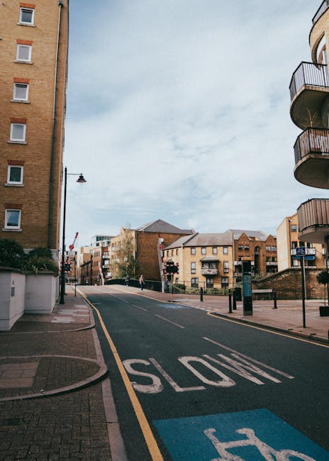 A view of a residential street in Brentford, with a slight incline and a clear sky overhead. On the left side, there is a tall, multi-storey brick residential building with several windows, while on the right side, a modern apartment building features curved balconies with metal railings. The street is lined with a sidewalk, and a black lamppost stands near the curb. The road has a prominent 'STOP' marking painted in white on the asphalt, and a blue parking zone marking is visible nearby. In the background, more residential buildings and trees can be seen, along with a few pedestrians walking along the sidewalk. The scene captures a calm urban environment suitable for house removals or furniture transport, with evidence of ongoing activity such as loading equipment or vehicles possibly nearby, as typical in home relocation processes. Man and Van Brentford often operate in this area for efficient packing and moving services.