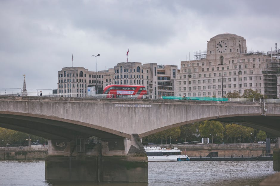 A view of Waterloo Bridge spanning the River Thames in London, with cloudy skies above. On the bridge, there is a red double-decker bus traveling across, and scaffolding surrounds part of a historic building with large clock faces on the right side. Below the bridge, the river features a moored white boat, and in the foreground, the water reflects the structure and boat. This scene captures the urban landscape, transportation, and architecture typical of central London, relevant to articles about home relocation or moving logistics in the Brentford area, such as those covered by Man and Van Brentford on their removals services page.