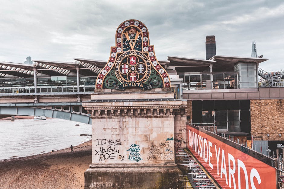 The image depicts the exterior of Kew Bridge in Brentford, featuring the iconic bridge structure with a decorative crest displaying coats of arms and the year 1862, mounted above the entrance. The bridge's stone supports are visible, with some graffiti and tags on the lower sections. To the right, a modern metal and glass building with a roof terrace overlooks the Thames River, which appears on the left side of the image with a partially visible boat. A large red and white banner with the text 'Bankside Yards' hangs along a construction fence on the right side, indicating ongoing development or renovation work nearby. The scene is set during daylight with overcast skies, and the environment suggests an urban riverside location suitable for home relocation activities involving furniture transport and packing. This image illustrates the type of scenic river views and structural elements relevant to house removals around riverside flats in Brentford, as referenced by Man and Van Brentford for efficient loading, transport, and moving logistics.