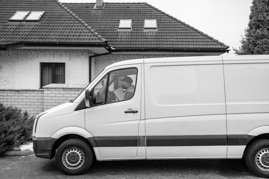 A white commercial van with a black stripe along its side is parked on the street in front of a residential property with a pitched tiled roof and a brick-and-finish exterior wall. The van's driver, w