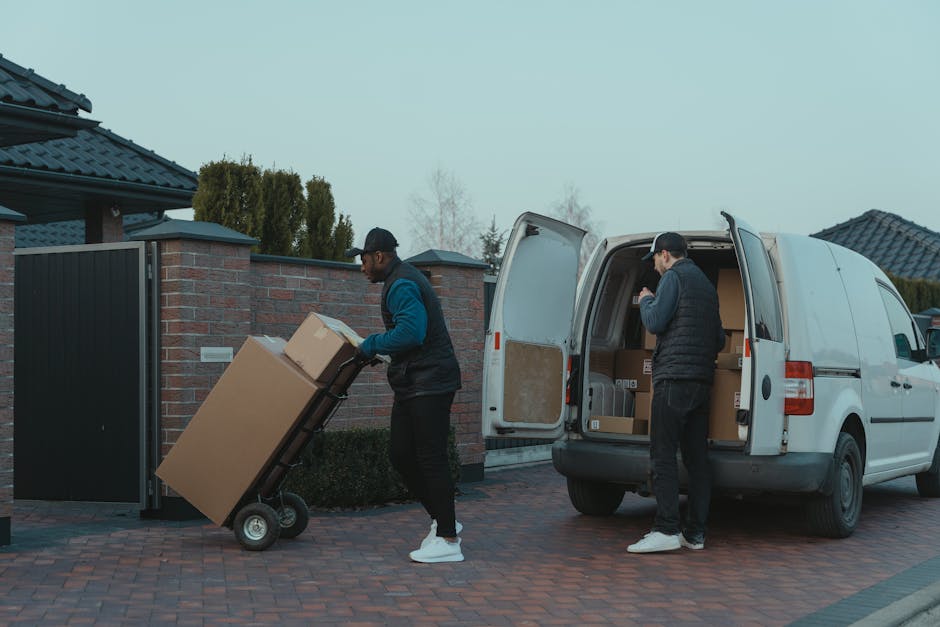 A white commercial van with a black stripe along its side is parked on the street in front of a residential property with a pitched tiled roof and a brick-and-finish exterior wall. The van's driver, w