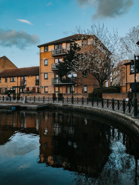 A multi-storey brick residential building with several balconies, situated behind a canal with a curved stone walkway and black metal railings along its edge. In the foreground, a man dressed in dark clothing is walking on the pavement, adjacent to a lamppost with multiple round white lights and a large tree with budding branches. Inside the property, partially visible through an open doorway, is a moving trolley loaded with cardboard boxes and wrapped furniture, supported by visible packing materials such as plastic wrap and blankets. The scene suggests a home relocation or furniture transport process managed by Man and Van Brentford, with the outdoor environment characterized by clear weather and soft natural light, suitable for logistics operations related to removals in Brentford flats and fixes.