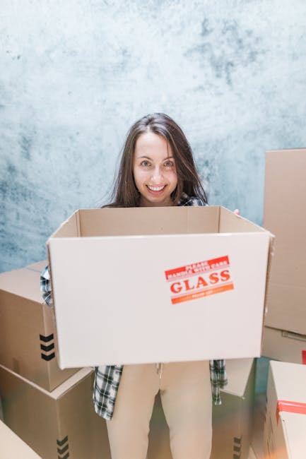 A young woman with long brown hair smiling while holding an empty cardboard box labeled 'Glass' during a home relocation process. She is surrounded by multiple other sealed cardboard boxes of various sizes, some with black and red tape, placed on a floor with a light blue textured wall in the background. The setting appears to be indoors, likely within a property preparing for a move, where packing and loading procedures are underway. The scene captures the typical elements of furniture transport, packing, and moving logistics, with the woman actively engaged in handling boxes as part of the removal process. This aligns with services offered by Man and Van Brentford, highlighting an efficient loading process involved in same-day removals and planning to avoid delays in Brentford.