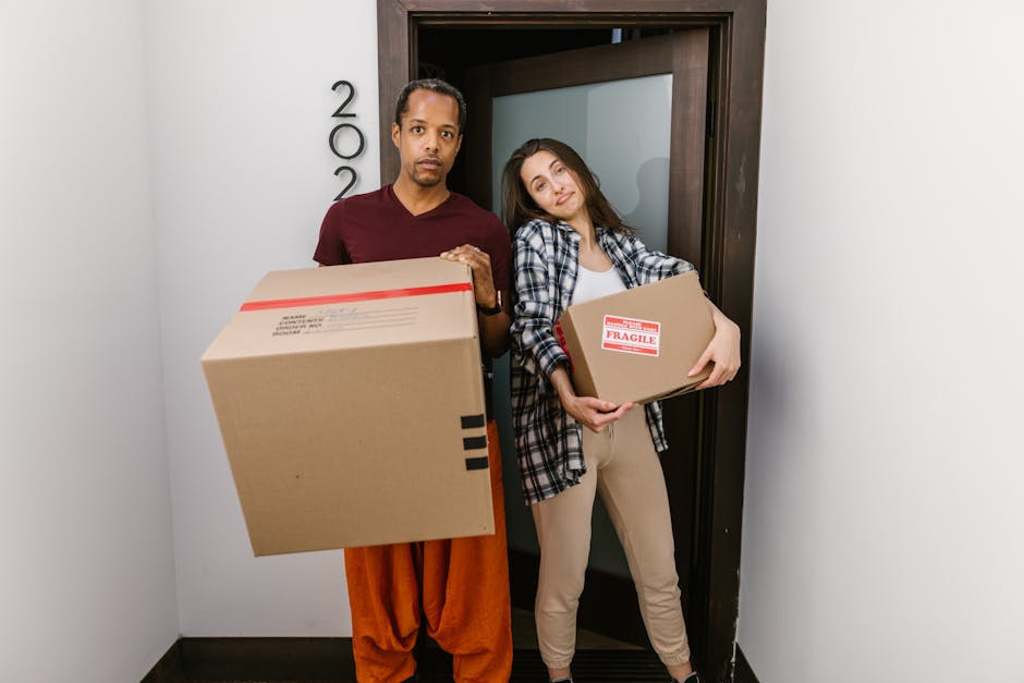 A man and a woman stand inside a doorway after completing a home relocation, both holding cardboard boxes with packing labels; the man holds a larger box, and the woman carries a smaller one labeled 'fragile.' They are positioned on a light-colored wall background with a dark wooden door frame behind them. The man wears an orange pair of trousers and a maroon t-shirt, while the woman is dressed in beige trousers and a plaid shirt over a white top. The scene takes place in an indoor hallway, adjacent to a small, plain white wall, with natural or artificial lighting illuminating the area. This image illustrates the loading process involved in furniture transport and packing during a same day removals, highlighting the careful handling of packages, which Man and Van Brentford offers as part of their house removal services, promoting efficient and careful home relocation logistics.