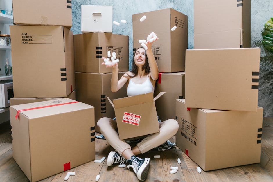 A young woman with long brown hair smiling while holding an empty cardboard box labeled 'Glass' during a home relocation process. She is surrounded by multiple other sealed cardboard boxes of various sizes, some with black and red tape, placed on a floor with a light blue textured wall in the background. The setting appears to be indoors, likely within a property preparing for a move, where packing and loading procedures are underway. The scene captures the typical elements of furniture transport, packing, and moving logistics, with the woman actively engaged in handling boxes as part of the removal process. This aligns with services offered by Man and Van Brentford, highlighting an efficient loading process involved in same-day removals and planning to avoid delays in Brentford.
