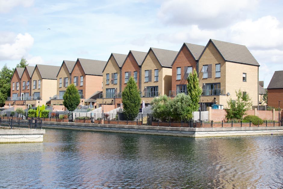 A row of modern, multi-storey residential houses with pitched roofs and brick exterior walls, situated along the edge of a calm canal or river. Each house has a small front garden enclosed by low fences, with a variety of trees and shrubs providing greenery. A paved pavement runs parallel to the water, and some houses feature exterior balconies and large windows. The sky is partly cloudy, and the overall scene suggests a peaceful suburban neighbourhood. The image, associated with house removals and relocation services by Man and Van Brentford, illustrates the type of homes near Brentford Lock that may require professional furniture transport, packing, and home relocation assistance. The setting emphasizes the importance of careful planning and logistics involved in moving in such residential areas, potentially involving the loading process of household belongings onto vehicles for transfer.