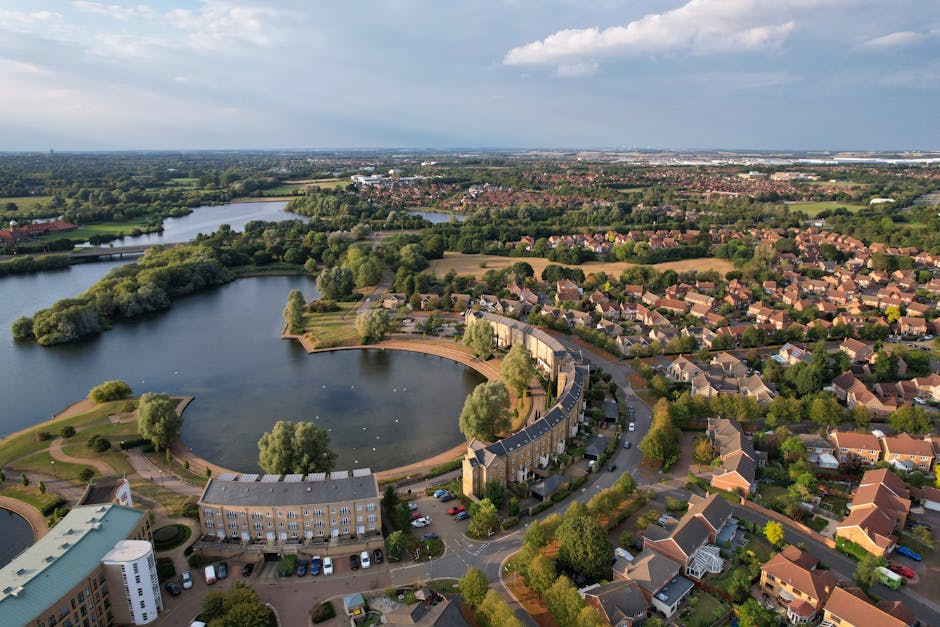 Aerial view of Syon Park near Brentford, showing a circular artificial lake surrounded by a curved residential building with a flat roof and light-colored exterior. The houseboats are moored along the edge of the lake, which is bordered by green grass and trees, creating a natural setting. To the right of the lake, rows of terraced houses with pitched roofs extend into the distance, with trees interspersed among the buildings. The scene captures the peaceful suburban environment, with parked cars along the streets, and a bridge crossing a river visible in the background. The sky is partly cloudy with sunlight illuminating the area, highlighting the landscaping and the various structures involved in a typical home relocation or furniture transport process. The presence of vehicles and outdoor furniture suggests ongoing or recent moving activities, supported by the professional removal services of Man and Van Brentford.