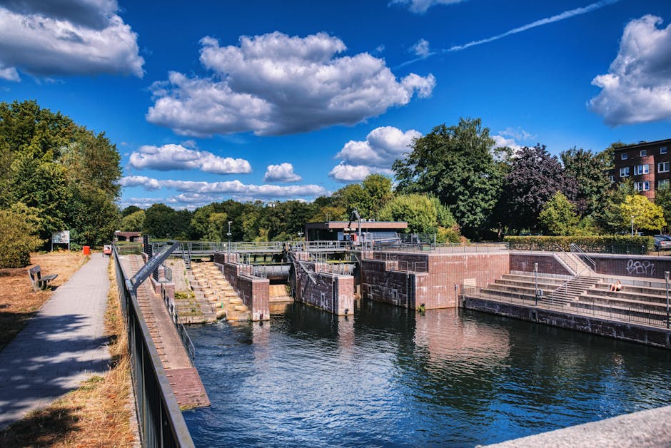 A row of modern, multi-storey residential houses with pitched roofs and brick exterior walls, situated along the edge of a calm canal or river. Each house has a small front garden enclosed by low fences, with a variety of trees and shrubs providing greenery. A paved pavement runs parallel to the water, and some houses feature exterior balconies and large windows. The sky is partly cloudy, and the overall scene suggests a peaceful suburban neighbourhood. The image, associated with house removals and relocation services by Man and Van Brentford, illustrates the type of homes near Brentford Lock that may require professional furniture transport, packing, and home relocation assistance. The setting emphasizes the importance of careful planning and logistics involved in moving in such residential areas, potentially involving the loading process of household belongings onto vehicles for transfer.