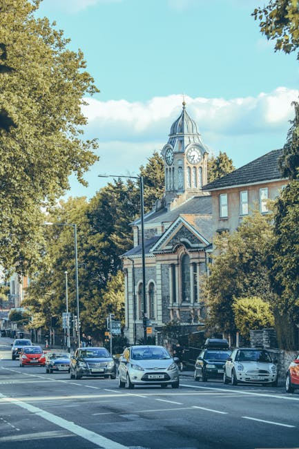 A street scene in Brentford showing several parked cars along the side of the road, including hatchbacks and sedans, with a large historic clock tower church visible in the background, surrounded by leafy green trees. The sky is blue with some scattered clouds, and the overall environment suggests a calm town centre setting during daytime. Although no interior or moving objects are visible, this scene could be part of the context for house removals or local relocation services offered by Man and Van Brentford, highlighting the typical environment where furniture transport and loading processes might take place during a home relocation or moving service in the TW8 area.